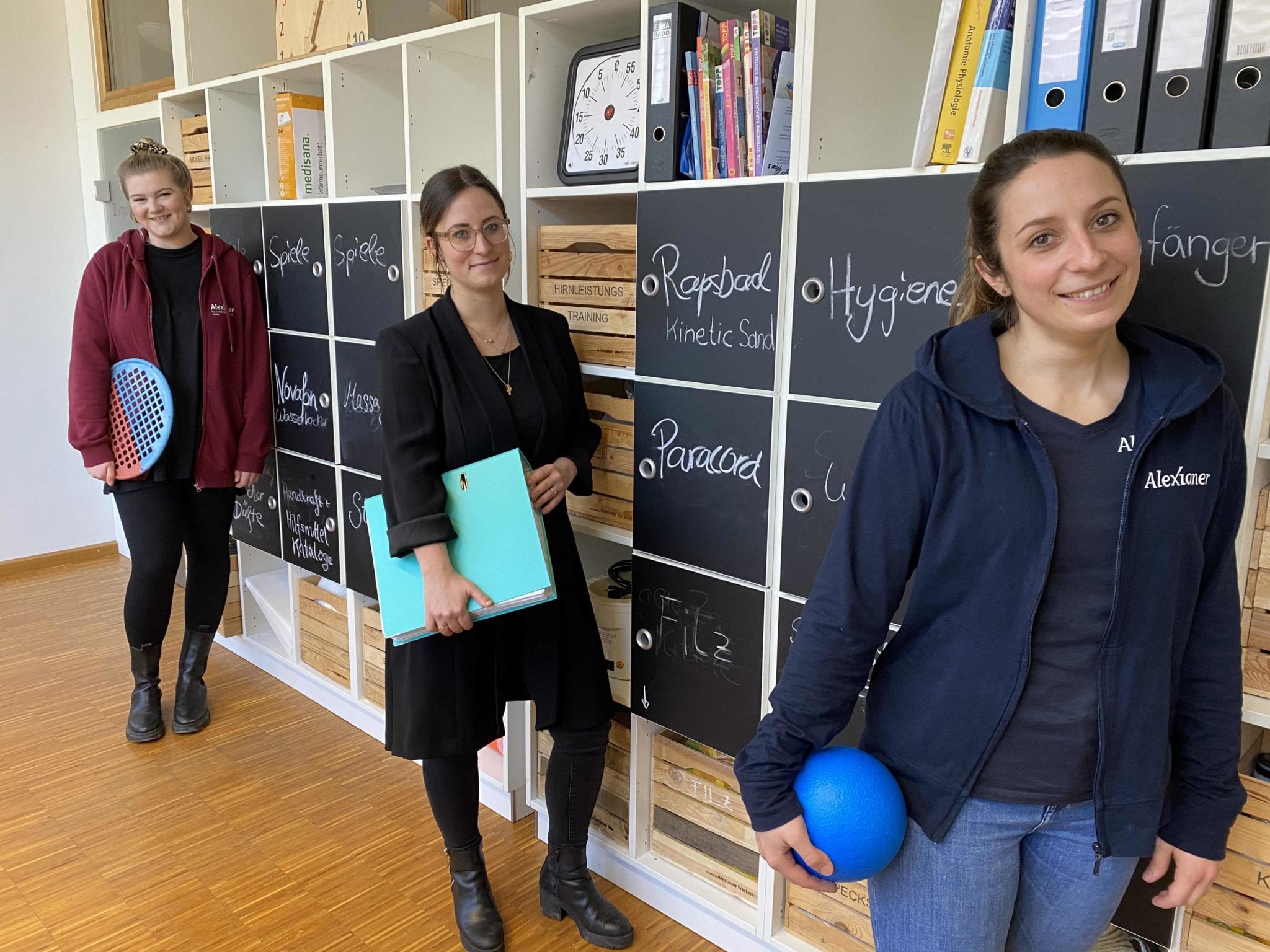 Ein Teil des Teams der Ergo- und Physiotherapie der Alexianer Werkstätten. Drei Mitarbeiterinnnen stehen in der Praxis vor einer Materialwand.